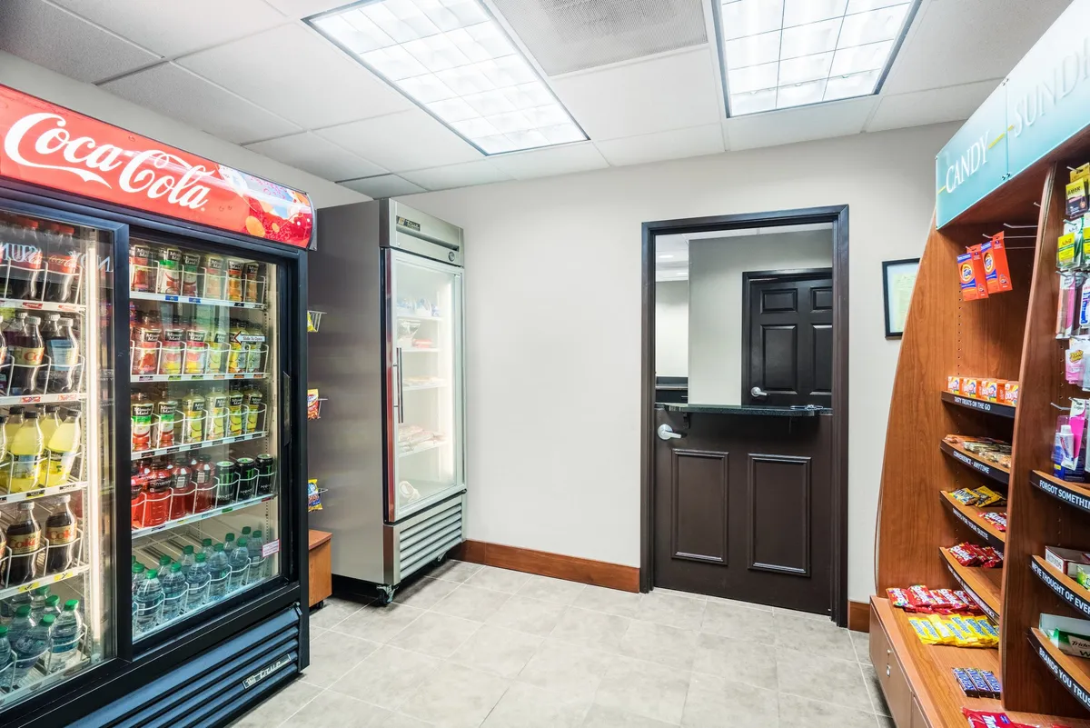 A cozy interior corner featuring a Coca-Cola vending machine, a snack shelf, and a refrigerator unit. The area is styled to encourage casual snacking, social interaction, or a quick refreshment break, emphasizing convenience for guests.