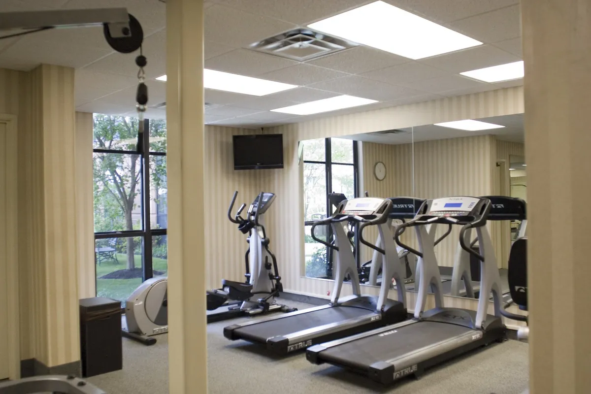 A modern, well-lit fitness center featuring treadmills, an elliptical machine, large windows for natural light, a television, and a wall clock. The space feels energetic and inviting, emphasizing both solo and community workouts.