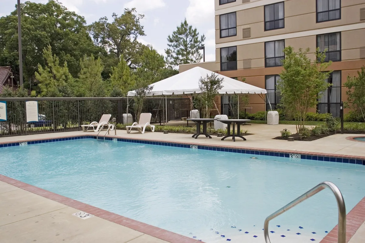 A welcoming exterior pool area, surrounded by lounge chairs, a tented space for gatherings, and lush trees. The setting transitions from serene mornings to lively sunset celebrations, suggesting an inviting community atmosphere and on-site relaxation options.