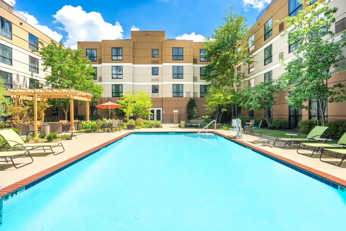 A bright and inviting exterior shot of the swimming pool area, surrounded by lounge chairs and a canopy-covered seating area. Lush greenery frames the space, and the scene suggests leisure, relaxation, and social gatherings. The pool is sparkling under the sun, providing a refreshing atmosphere.