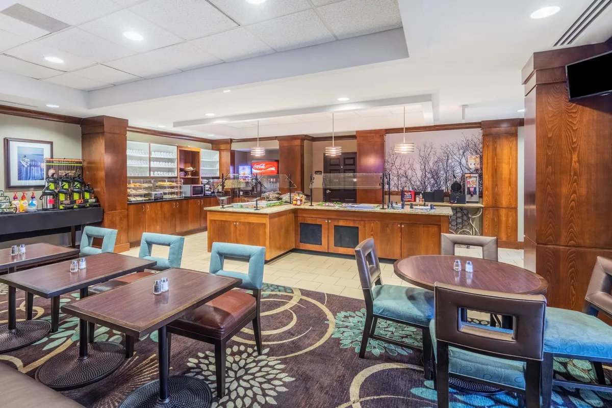 A bright, well-lit studio with a coffee station and a food counter, surrounded by tables and chairs where guests are socializing, enjoying breakfast, or working on laptops. Natural light streams in, illuminating an inviting space decorated for comfort and practicality, with ample seating that could double as a workspace.
